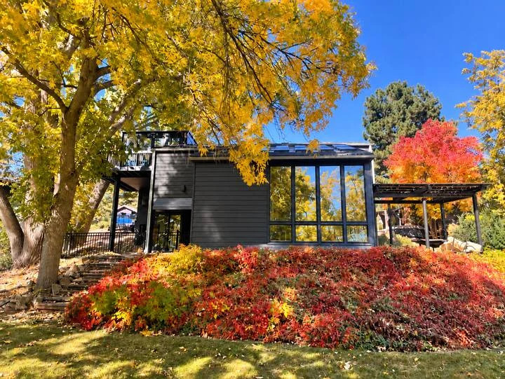 Backyard view of the residence with tall windows, dark siding, pergola and colorful autumn landscaping