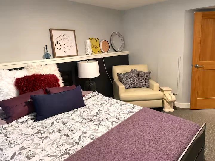 Ground-level bedroom with layered pillows, purple blanket, bedside lamp and cream armchair