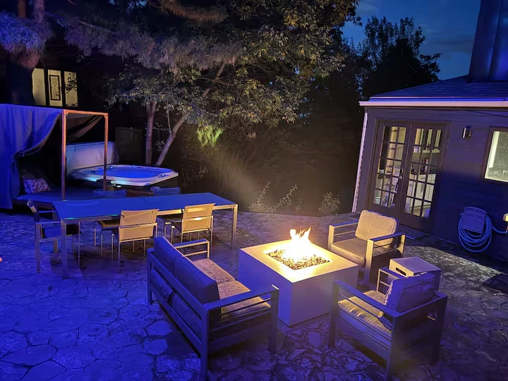 Courtyard lounge seating around a glowing fire pit with hot tub and trees behind at night