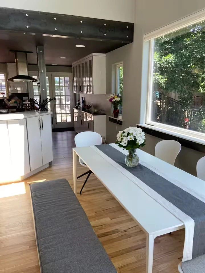 White dining table with flowers, chairs and upholstered bench beside a garden-facing window and kitchen