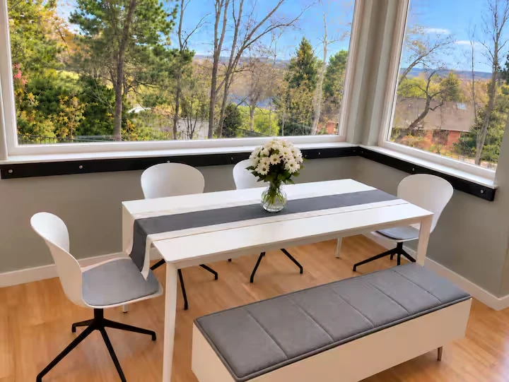 Breakfast nook with white table, chairs and gray bench surrounded by windows overlooking trees
