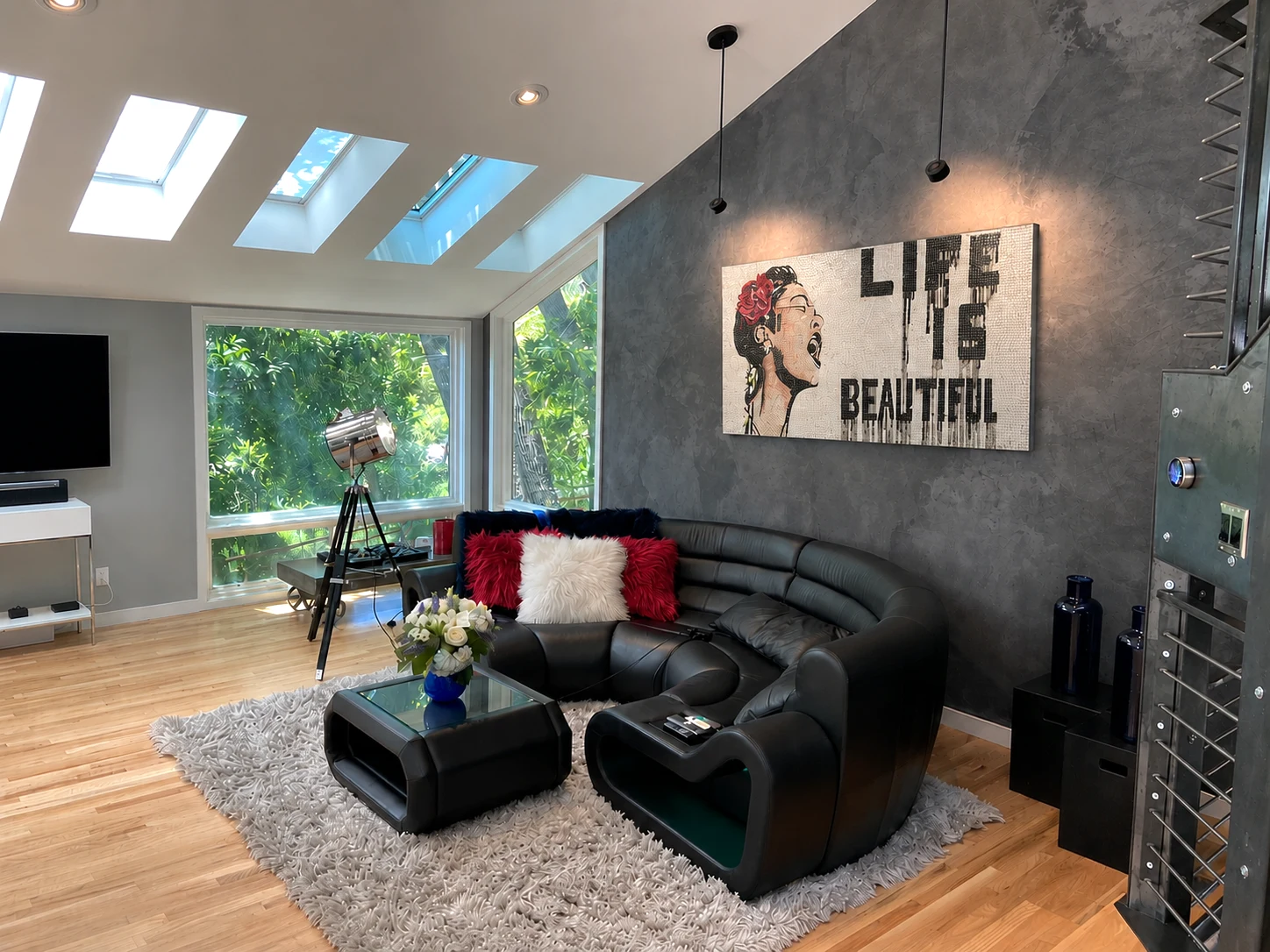 Curved black sofa beneath the Life Is Beautiful artwork, beside a tripod spotlight and steel staircase