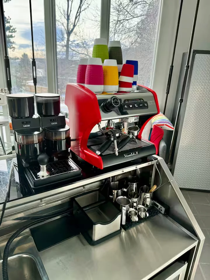 Red espresso machine with colorful cups, two coffee grinders and stainless steel preparation counter beside windows