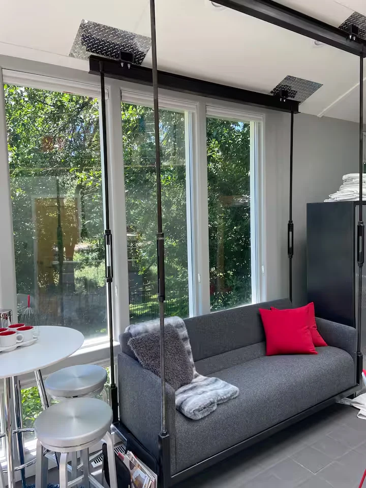 Suspended gray sofa with red cushion beside a round café table and garden-facing sunroom windows