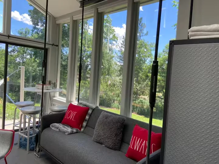 Floating gray sofa with red cushions suspended in a sunroom corner overlooking the lawn and trees